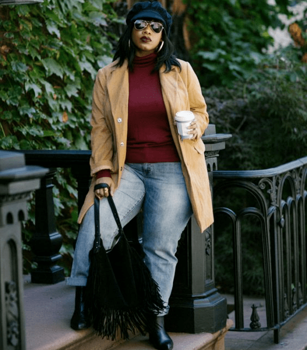 chica curvy morena usando un gorro negro, lentes de sol, top de manga larga y cuello alto color guinda, saco beige largo, jeans a la cintura, botines negros de piel, bolso negro de mano con flecos