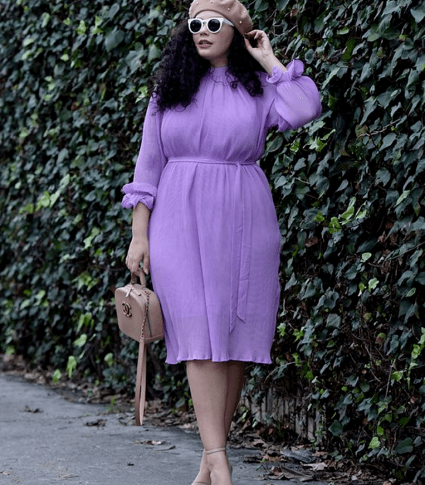 chica curvy de cabello oscuro chino usando un beret beige, lentes de sol blancos, vestido lila de manga larga, bolso beige de mano y sandalias de tacón beige