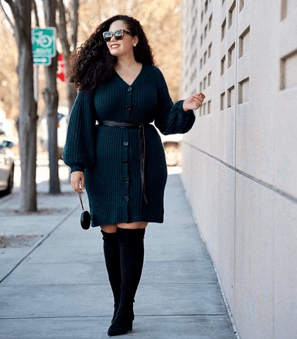 chica curvy de cabello chino largo usando lentes de sol, un vestido tejido de botones color azul marino con manga larga y cuello en V, cinturón negro en la cintura, botas largas negras con tacón y bolso mini negro de mano