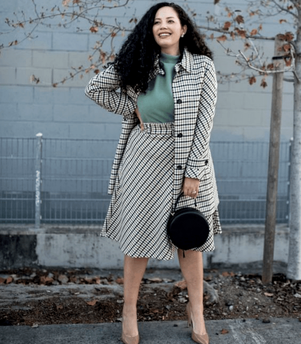 chica curvy de cabello chino largo usando una blusa color menta de cuello alto, saco largo beige con líneas negras, falda a la cintura larga color beige con líneas negras, bolso negro de mano y zapatos de tacón color beige