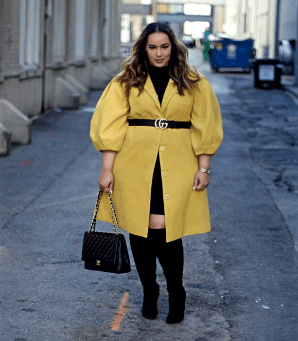 chica curvy de cabello claro usando un top negro de cuello alto, vestido amarillo de mangas abombadas, cuello en V, cinturón negro a la cintura y falda negra, botas largas de tacón y bolso negro de mano