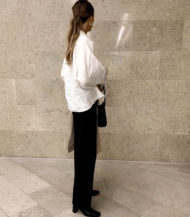 chica rubia con camisa oversized blanca, pantalones de vestir negros, botines negros de tacón y bolso negro de mano