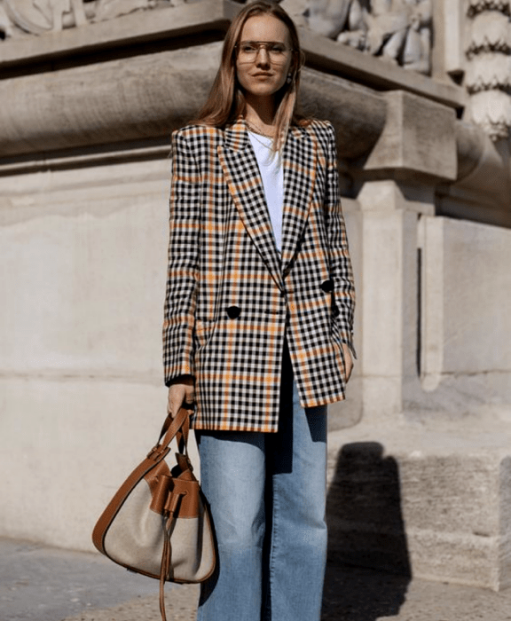 chica rubia usando lentes, camiseta blanca, blazer oversized amarillo con negro de cuadros, jeans holgados y bolso beige y café de piel