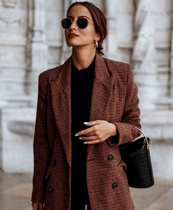 chica de cabello castaño usando lentes de sol, top negro, blazer café de cuadros y jeans negros, bolso bucketbag negro de piel