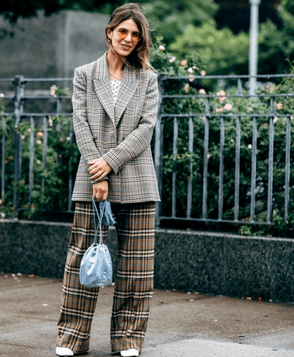 chica de cabello claro usando lentes de sol, top blanco con lunares, blazer gris de cuadros, pantalones de cuadros cafés de vestir, botines blancos, bolso bucketbag celeste