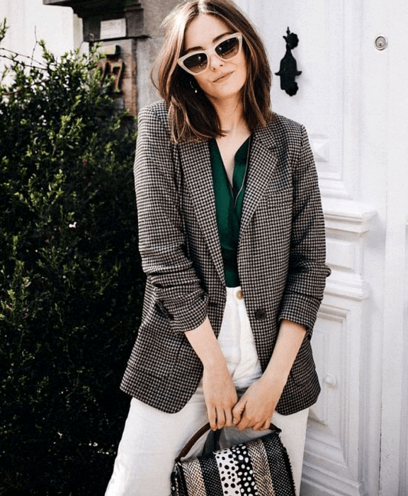 chica de cabello castaño claro usando lentes de sol beige, blusa verde esmeralda, blazer de cuadros café con gris, pantalón blanco, bolso de mano estampado blanco, gris y beige