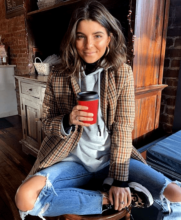 chica de cabello claro usando una sudadera gris, blazer café de cuadros, jeans rotos en las rodillas, tenis deportivos café con negro y blanco