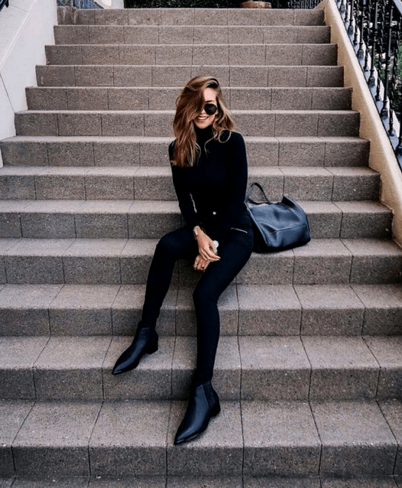 chica de cabello claro usando lentes de sol, top de manga larga y cuello alto negro, leggings negros y botines de cuero sin tacón, bolso de mano  negro