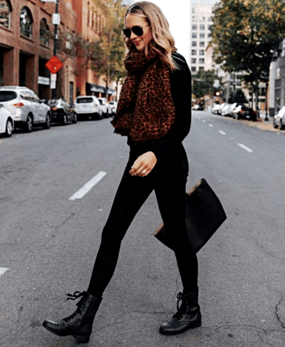 chica rubia usando lentes de sol, bufanda café de animal print, suéter negro de manga larga, leggings negros y botines negros sin tacón y bolso de mano  negro