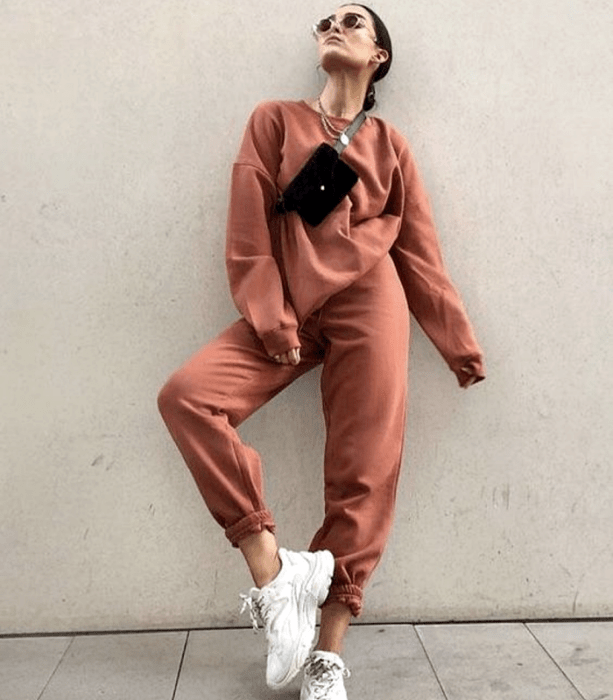 chica de cabello oscuro usando lentes de sol, sudadera oversized, pants naranjas y tenis blancos deportivos y bolso negro