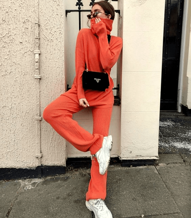 chica de cabello castaño usando lentes de sol, sudadera de cuello alto color naranja, bolso negro de mano, pants holgados naranjas y tenis blancos