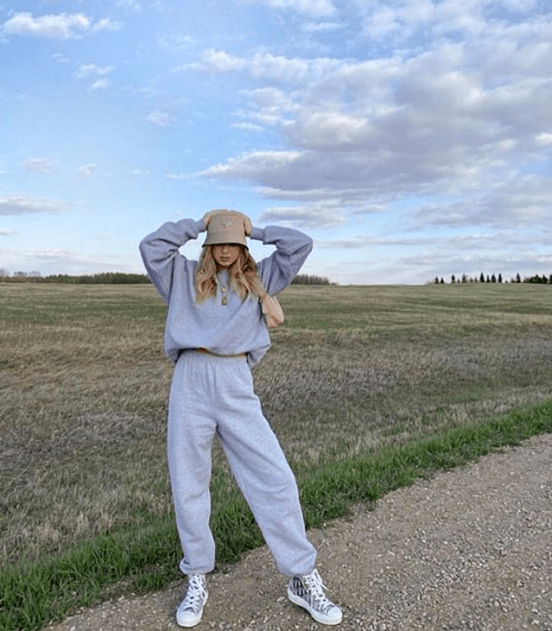 chica rubia con un bucket hat beige, sudadera oversized gris, pants deportivos grises, tenis converse blancos con negro y bolso de mano beige