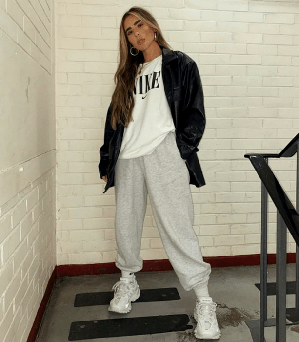 chica de cabello claro largo usando una camiseta blanca oversized, chamarra de cuero, pants grises y tenis blancos deportivos