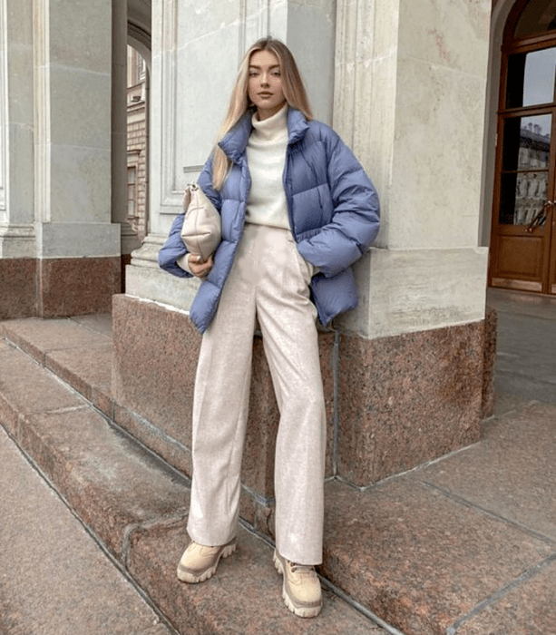 chica rubia usando un suéter blanco de cuello alto, pants beige holgados, chamarra puffer jacket azul celeste, bolso de mano beige y botas beige de suela gruesa