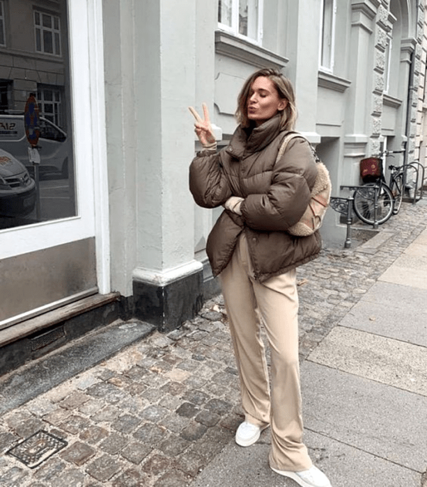 chica rubia de cabello corto usando una chamarra puffer jacket café, bolso de mano beige, pants beige holgados y tenis blancos