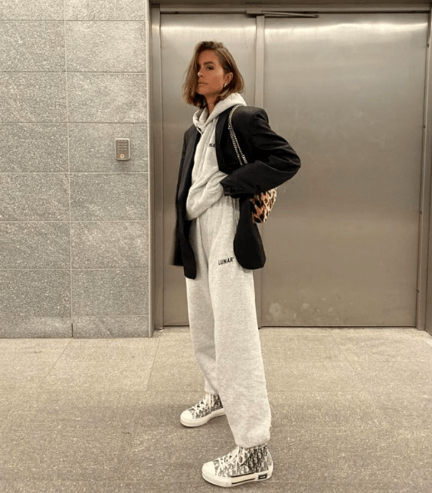 chica de cabello corto usando una sudadera gris, blazer negro, pants grises holgados, tenis converse blancos con negro y bolso de mano de animal print