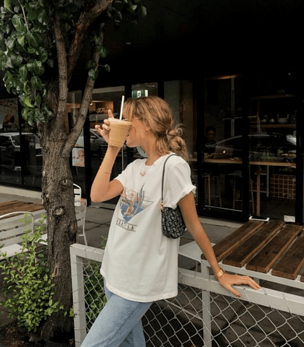 chica rubia usando lentes, camiseta blanca estampada, jeans holgados y bolso de mano  negro con blanco