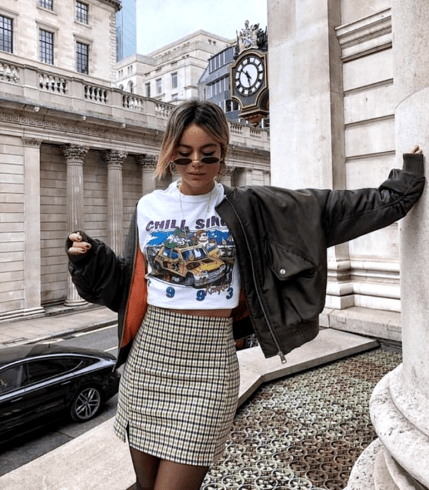 chica de cabello castaño claro usando lentes de sol, camiseta blanca gráfica estampada, minifalda beige de cuadros, medias negras y chamarra de cuero negra con forro naranja