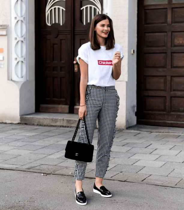 chica de cabello castaño usando una camiseta blanca gráfica estampada, pantalones de vestir blancos de cuadros negros, falts negros de suela blanca y bolso de mano negro