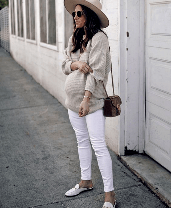 chica de cabello largo embarazada usando lentes de sol, sombrero de ala grande beige, suéter oversized beige, skinny jeans blancos, zapatos blancos loafers, bolso café crossover