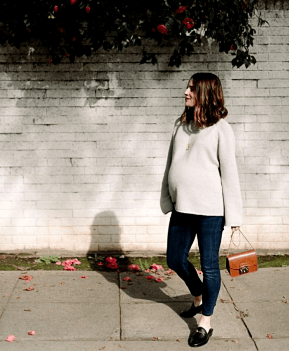 chica de cabello castaño, embarazada usando un suéter oversized gris, skinny jeans, zapatos negros tipo loafers, bolso de mano naranja