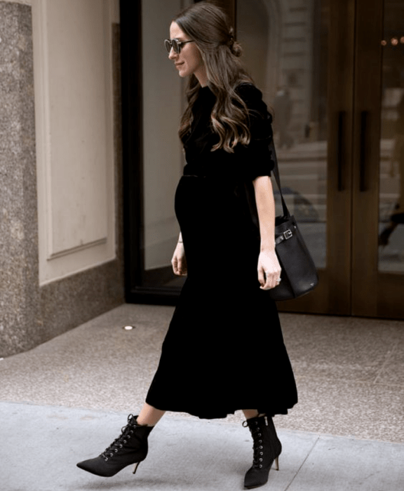 chica embarazada de cabello castaño largo usando lentes de sol, un vestido maxi negro de mangas 3/4 y botines de tacón 