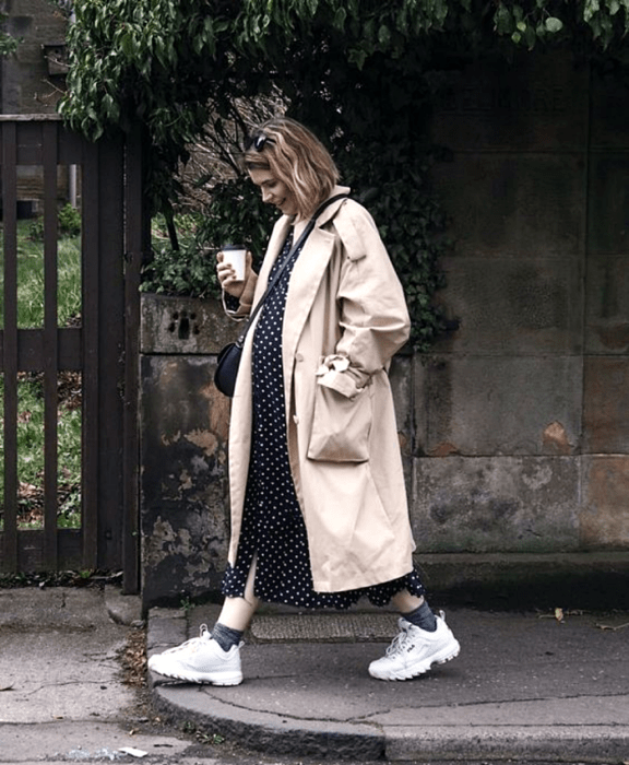 chica de cabello corto embarazada usando un vestido largo negro con lunares blancos, abrigo largo beige, tenis blancos, bolso negro crossbody bag