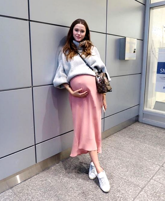 chica de cabello castaño, embarazada, usando un suéter gris oversized, falda larga rosa, tenis blancos y bolso crossover café