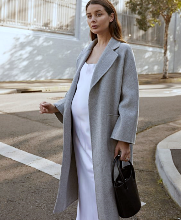 chica de cabello castaño embarazada con vestido de satén blanco, abrigo largo gris y bolso negro bucketbag