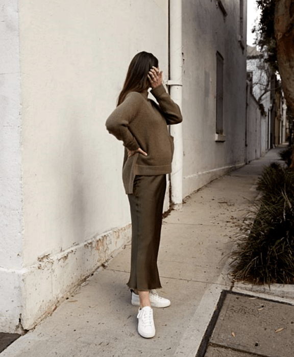 chica de cabello largo embarazada usando un suéter oversized verde oscuro, una falda de satén verde oscuro y tenis blancos