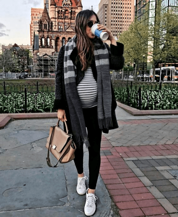 chica de cabello castaño embarazada, usando lentes de sol, bufanda gris, chamarra negra afelpada, top de rayas blancas con negro, leggings negros, tenis deportivos y bolso de mano beige