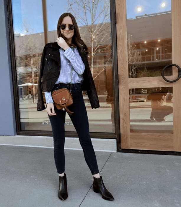 chica de cabello castaño usando lentes de sol, camisa azul de rayas blancas, chaqueta de cuero, skinny jeans negros, botines negros de tacón y bolso café