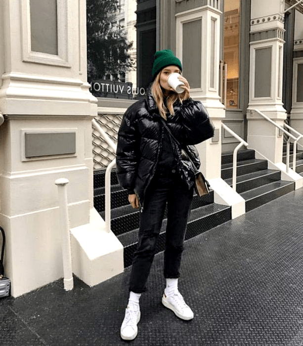 chica de cabello rubio usando un gorro verde, chamarra puffer jacket negra, skinny jeans negros, tenis deportivos blancos y bolso negro con café