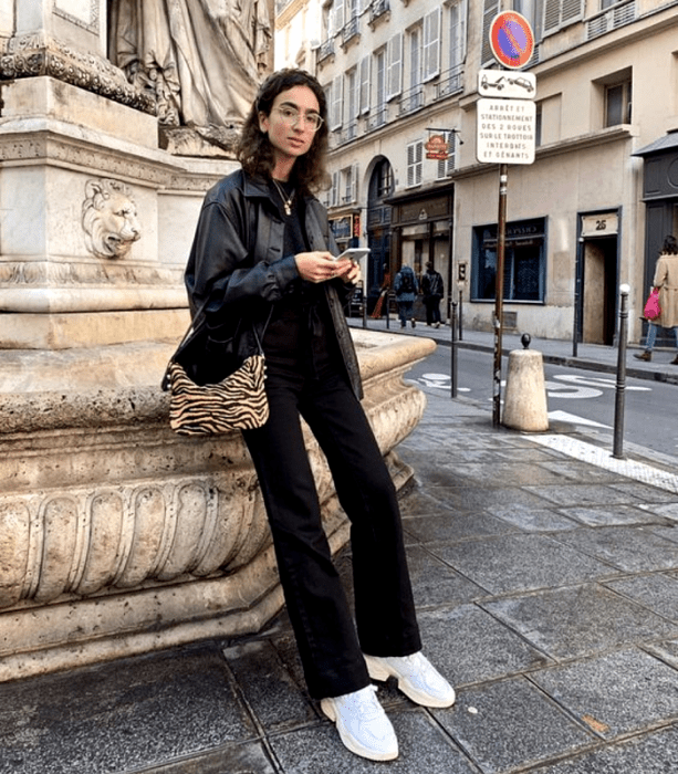 chica de cabello claro usando lentes, top negro, chaqueta de cuero, bolso de mano de animal print, jeans negros acampanados, tenis deportivos blancos