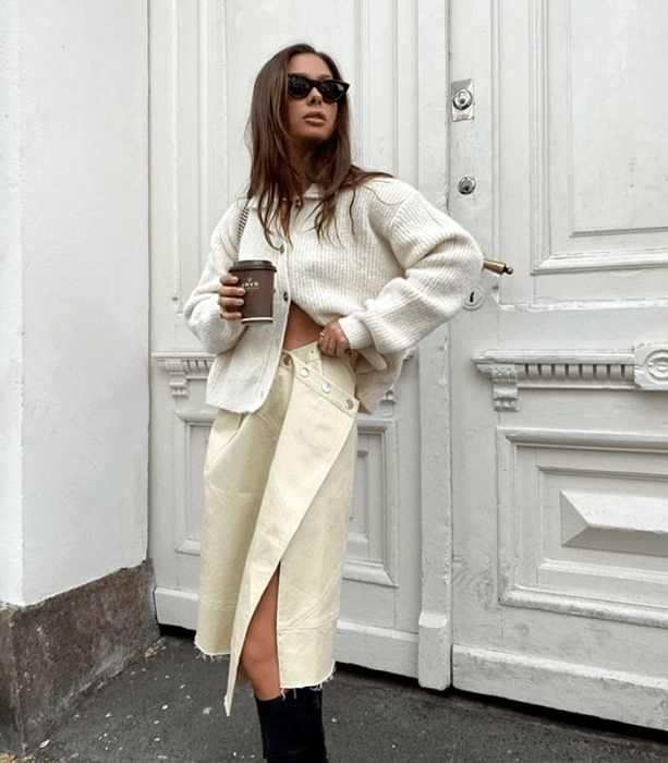 chica de cabello castaño usando lentes de sol, cárdigan oversized blanco, falda de cuero beige, botas largas negras