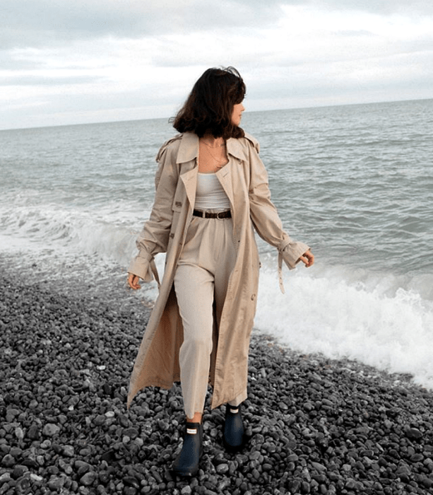 chica de cabello oscuro usando un abrigo largo beige, top blanco, cinturón negro, pantalones de vestir beige, botines negros de lluvia