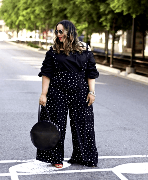 chica curvy de cabello claro usando lentes de sol, blusa sin hombros de manga 3/4, jumpsuit negro de lunares blancos y tirantes, bolso de mano negro redondo y sandalias de tacón rojas