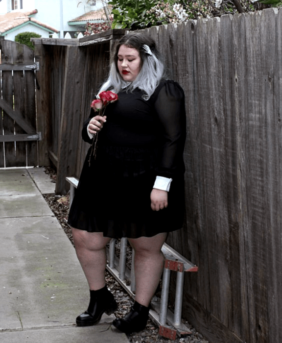 chica curvy de cabello gris usando un vestido negro de manga larga y puños blancos, medias negras semitransparentes y botines negros de tacón