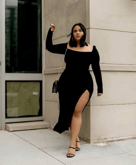 chica morena curvy usando un vestido negro ajustado de manga larga y escote cuadrado con apertura en la pierna y bolso negro pequeño