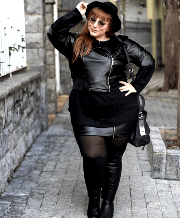 chica curvy pelirroja usando un sombrero negro, lentes de sol, chamarra de cuero negra, top negro largo, minifalda negra, medias negras, botas largas negras de tacón y bolso negro