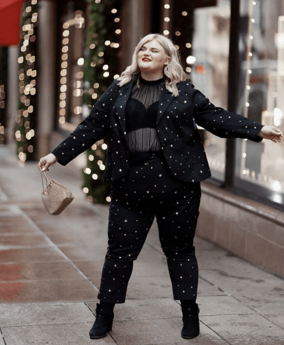 chica curvy rubia usando un top semitransparente negro, bralette negro, saco negro con estrellas doradas, pantalón de vestir negro con estrellas doradas, botines negros de tacón y bolso mini de mano beige