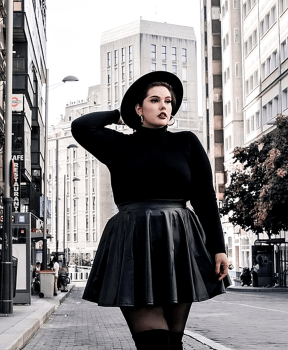 chica curvy de cabello negro usando un sombrero negro de ala ancha, suéter negro de cuello alto y manga larga, falda de piel negra con tablones, medias negras y botas largas negras