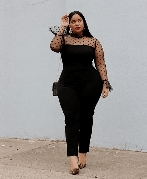 chica curvy morena de pelo largo, usando un jumpsuit negro con mangas y hombros semitransparentes con lunares negros, tacones de animal print y bolso negro mini