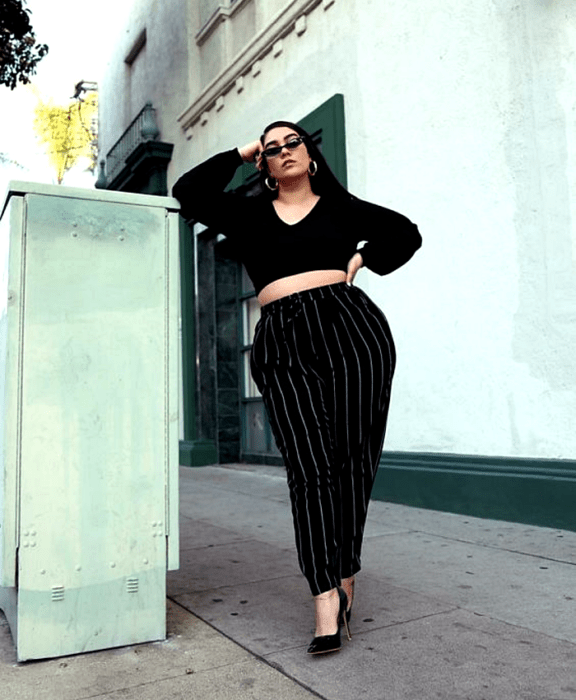 chica curvy de cabello largo oscuro usando lentes de sol, blusa crop top negra de mangas abombadas, pantalones negros de rayas blancas y tacones negros