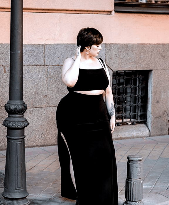 chica curvy de cabello corto negro, usando un crop top negro de tirantes delgados, falda maxi negra con abertura en la pierna y bolso negro