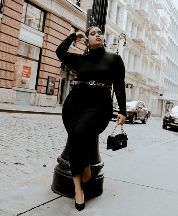 chica curvy morena usando un vestido tejido maxi negro de manga larga y cuello alto, cinturón café y bolso negro de mano, zapatos de tacón