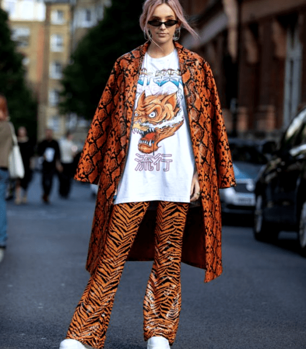 chica rubia usando lentes de sol, camiseta blanca oversized con estampado de dragon, abrigo largo de cuero naranja con líneas negras, pantalones de cuero acampanados color naranja con animal print de tigre y tenis blancos