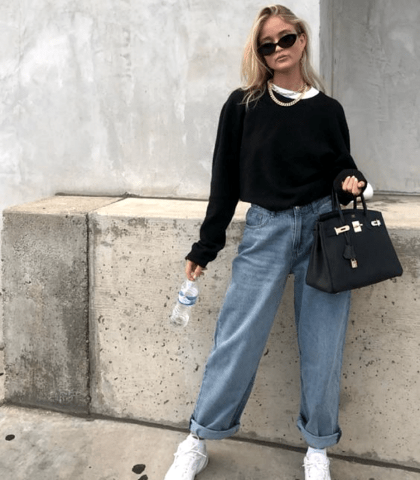 chica rubia usando lentes de sol, sudadera negra, camiseta blanca, jeans holgados y tenis blancos,  bolso  negro de mano