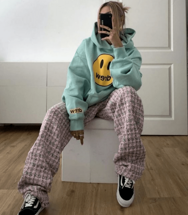 chica rubia usando una sudadera oversized azul celeste con estampado de carita feliz amarilla, pantalones holgados rosas con cuadros grises y tenis negros vans