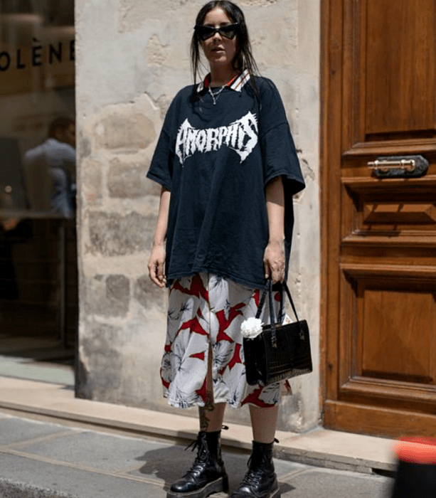 chica de cabello oscuro usando lentes de sol, camiseta oversized azul marino con estampado blanco, vestido blanco largo de flores blancas con rojo, botas negras de suela gruesa y bolso negro de mano